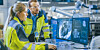 Two people working in a factory control robots from a workstation
