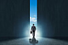 Business suit wearing man walks out of closing door in darkened room into the bright sunlight and blue sky