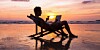 Someone using their laptop in a deck chair on the beach, in the sea, as the sunsets while holding a martini
