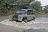A convoy of classic Land Rover vehicles, also known as Landy, crosses a shallow river in a tropical outdoor setting,