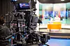 Studio camera recording a news anchor at an illuminated desk in a TV newsroom.