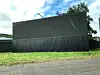 Rear view of a large dark billboard beside a corrugated fence, grassy verge and road under a partly cloudy sky.