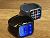 Two black smartwatches on a wooden surface, one showing a blue health screen and the other app icons.