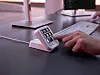 A hand presses a small backlit keypad beside a computer keyboard on a desk.